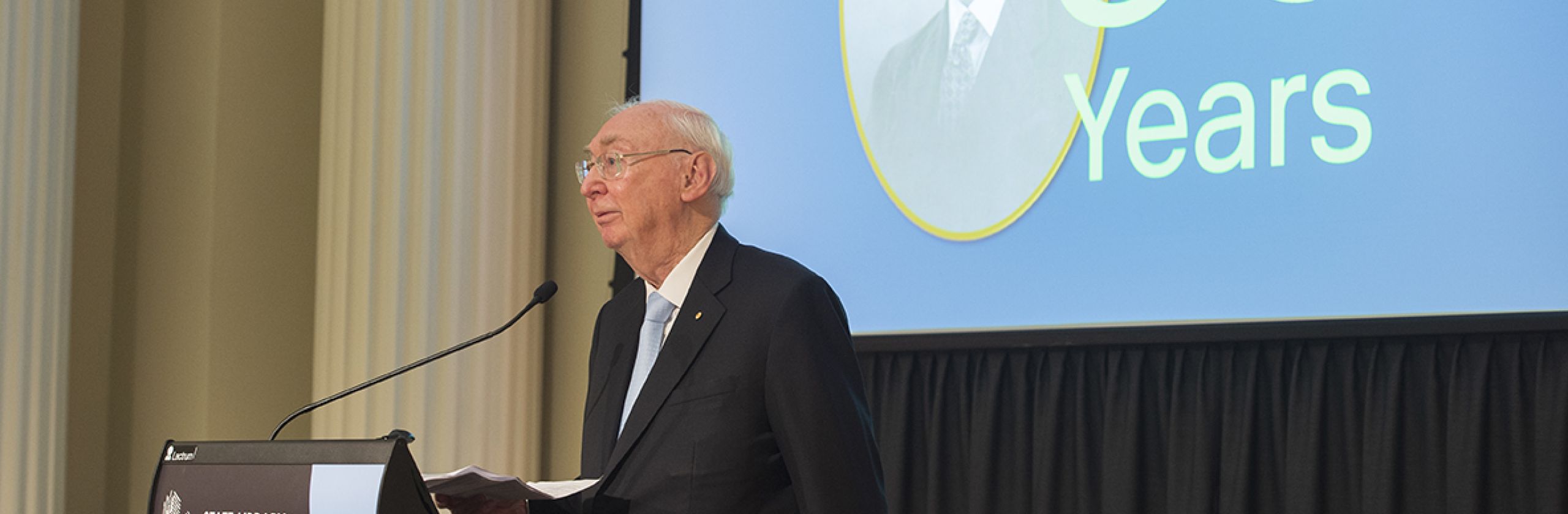 Charles Goode, AC, Chairman of The George Alexander Foundation speaking at a podium. In the background a large screen displays an image of George Alexander and '50 Years'.