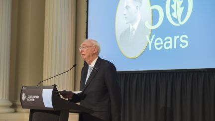 Charles Goode, AC, Chairman of The George Alexander Foundation speaking at a podium. In the background a large screen displays an image of George Alexander and '50 Years'.