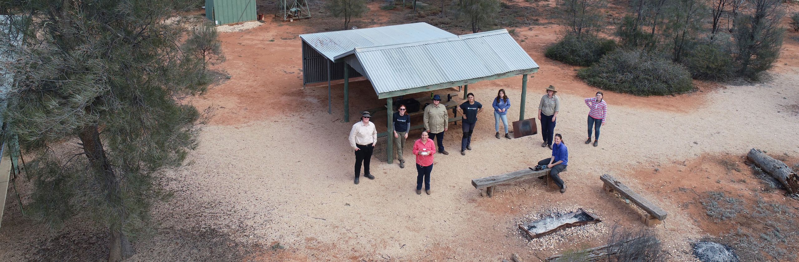 A birds eye view of Earthwatch fellows standing in bushland near a hut with a corrugated roof..