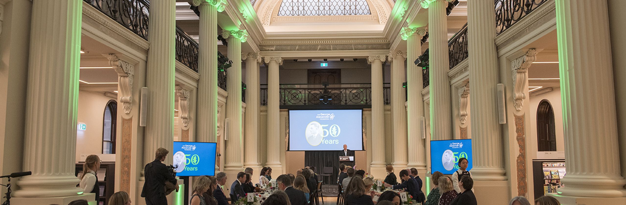 Guest at teh dinner seated at rectangular tables down the length of the Queen's Hall with large screens at the end.