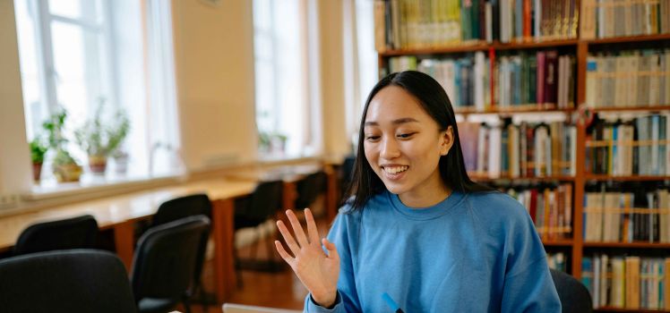 A young woman in a blue sweatshirt waves to her laptop. She is sitting at a desk with a bookcase behind her. 