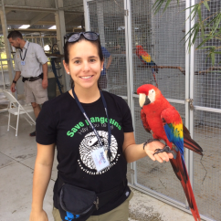 Bianca Papadopoulos, ISSI fellow. Woman with a large red, yellow and blue bird perched on her hand standing in front of an animal enclosure.