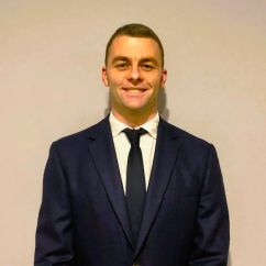 Man in dark suit and tie smiling, standing in front of a white wall.