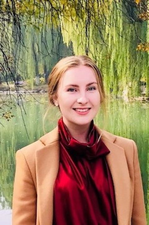 Georgia McDonald, Griffith University Scholar. Young woman wearing a red top smiling and standing in front of a lake.