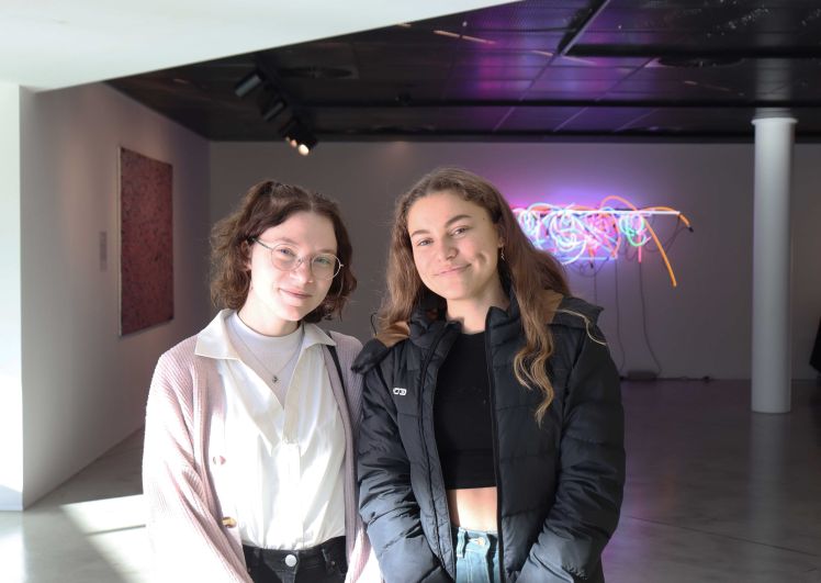 Two young women, Murdoch University scholars, smiling at an indoor event. Two pieces of artwork are displayed in the background.