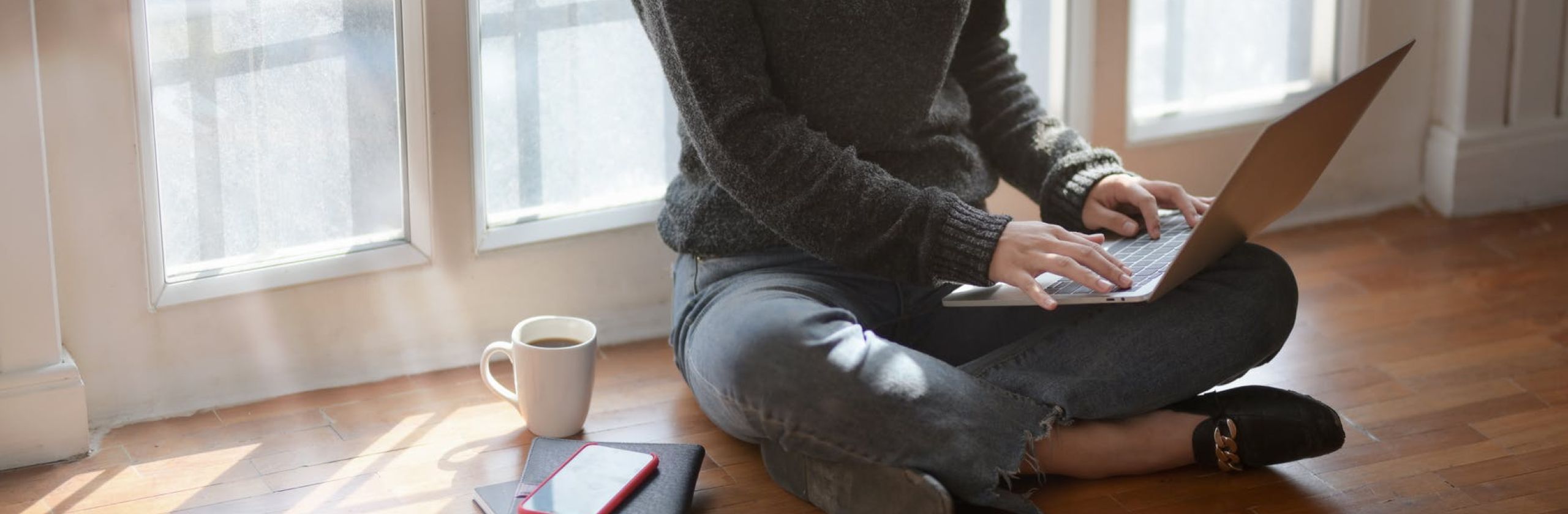 person sitting on wooden floors studying on a laptop. A mug and notebooks positioned next to them