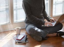 person sitting on wooden floors studying on a laptop. A mug and notebooks positioned next to them