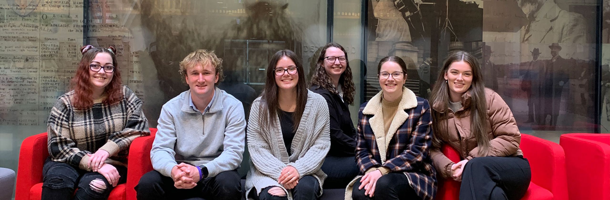 A group of Deakin University scholars sitting on red chairs in front of an image of Alfred Deakin.