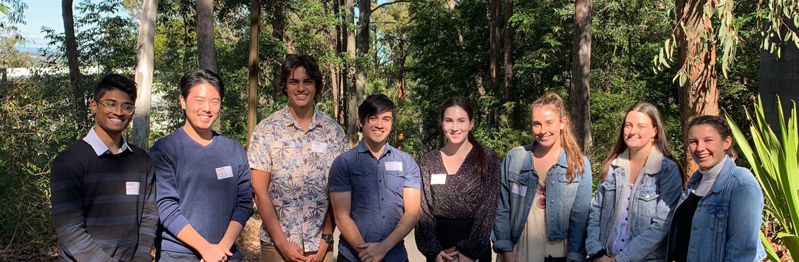A group of eight young students smiling and standing in a semi-circle outside.