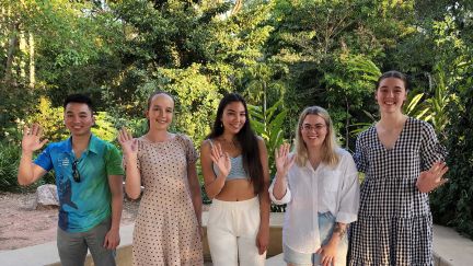 Five James Cook University scholars waving, standing outside with trees and plants in the background.