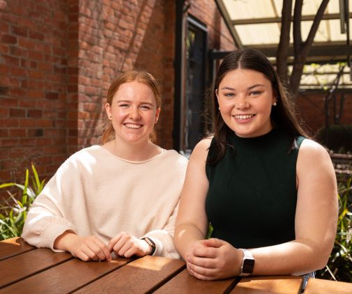 Two UniSA Scholars smiling and sitting outside at a wooden table. 
