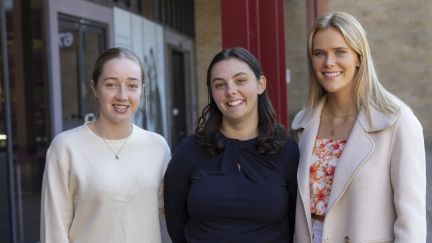 Three university students standing together and smiling. One woman has brunette hair in a bun and is wearing a cream jumper. Another woman has long dark brown hair and wears a navy jumper. 