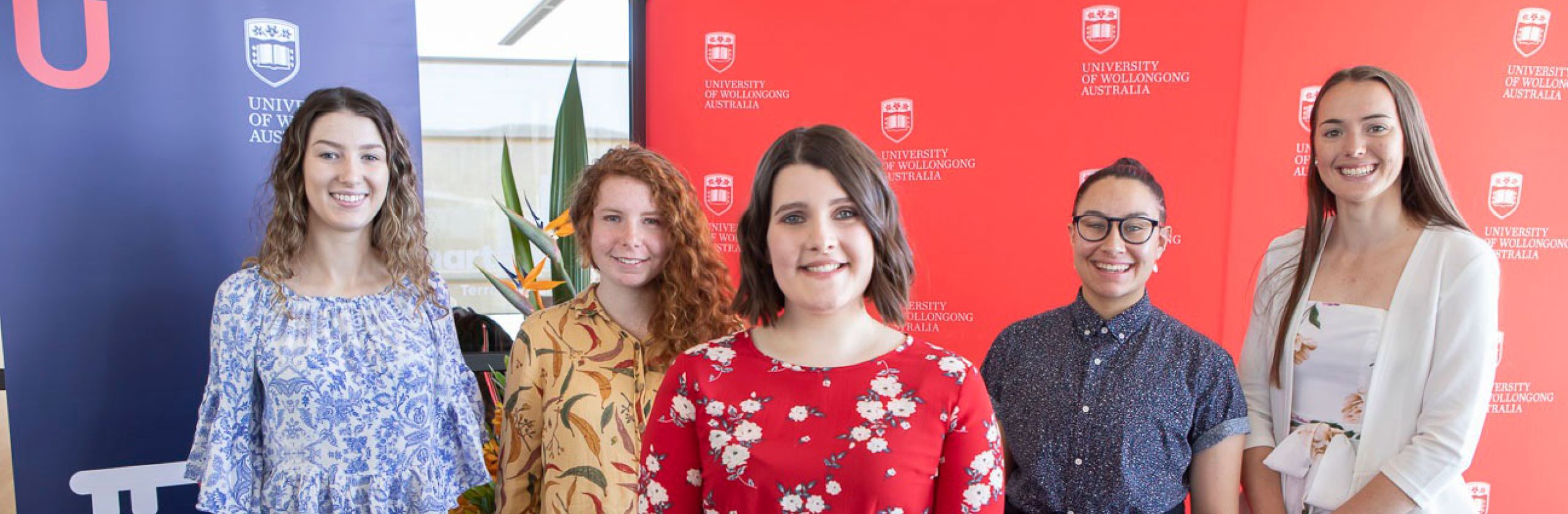 Five University of Wollongong Scholars standing in a triangle formation and smiling in front of a red and blue media wall.