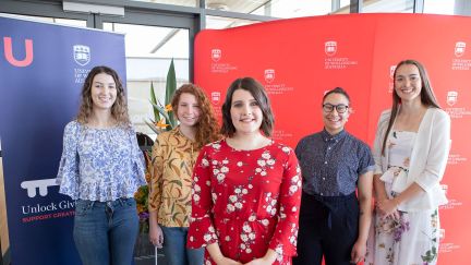 Five University of Wollongong Scholars standing in a triangle formation and smiling in front of a red and blue media wall.