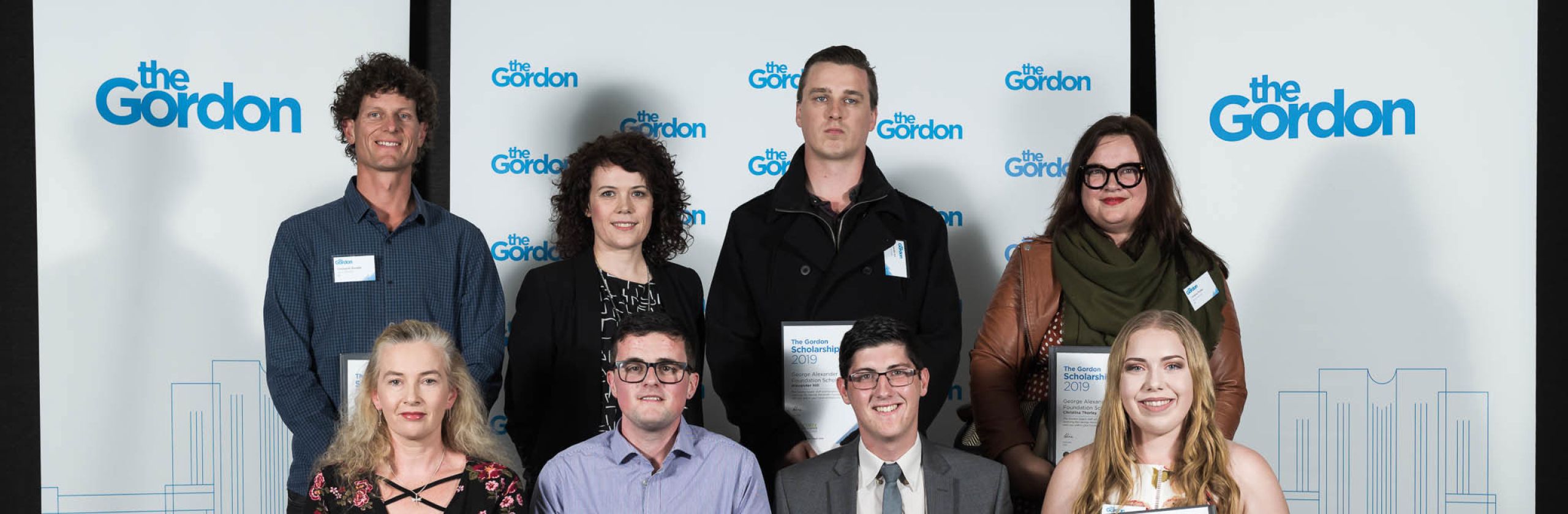 The Gordon scholars holding scholarship certificates in front of a banner with The Gordon logo. 