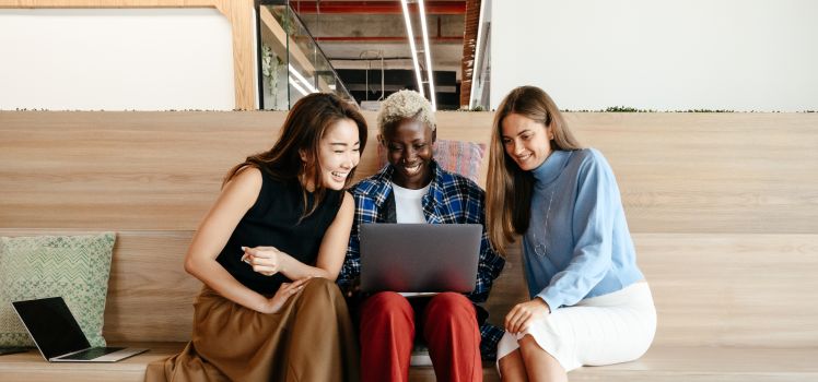 three women siting on a bench smiling at a laptop.