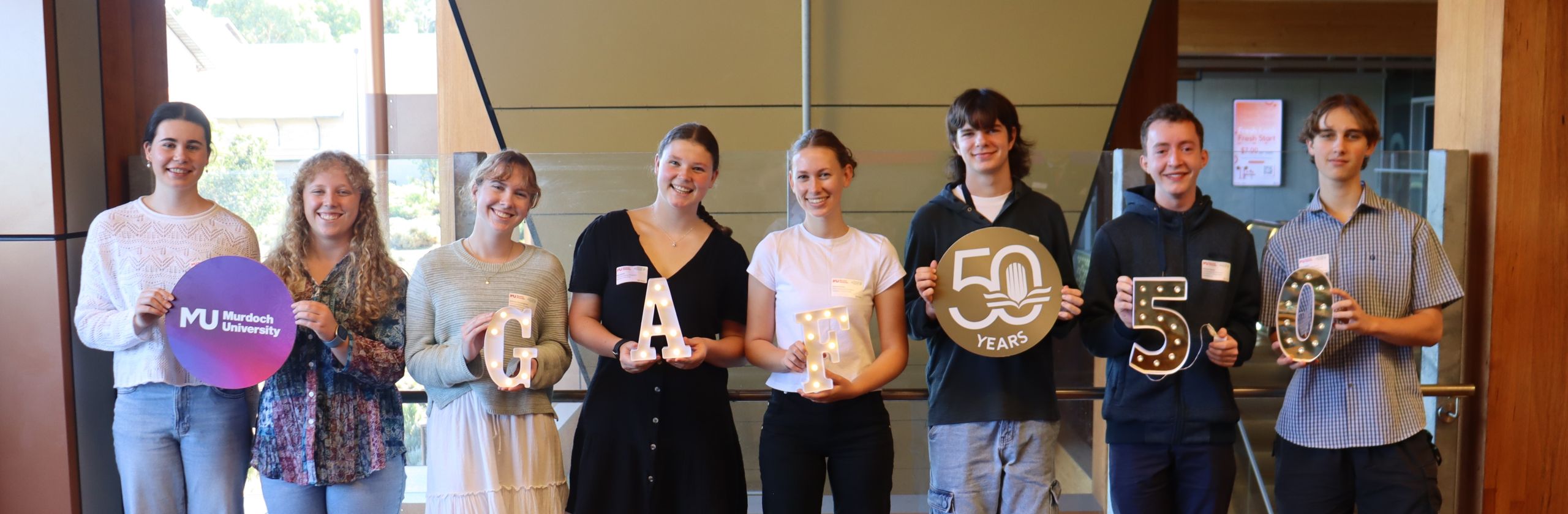 Eight yougn univeristy students standign in a line facing the camera holding signs to celebrate Murdoch Unviersity's 50th anniversary.