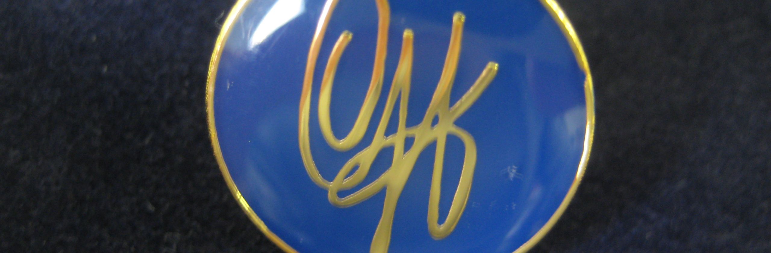Circular pin embossed in blue with gold signature and edging