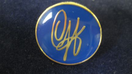 Circular pin embossed in blue with gold signature and edging